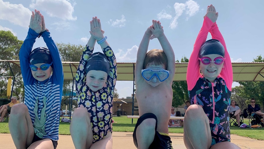 four children with goggles kneel on edge of pool with hands above head ready to dive in.