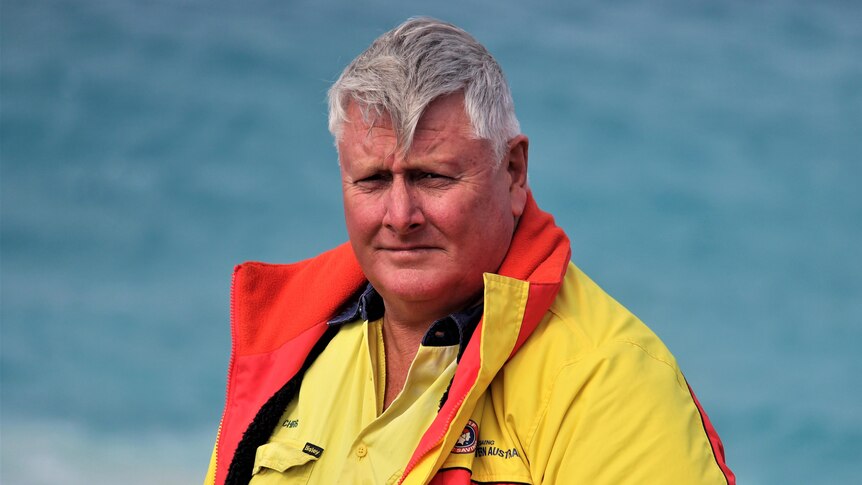 A serious-looking heavy man with grey hair, wears a surf life saving jacket and stands near the ocean.