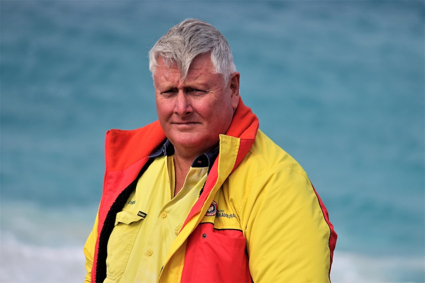 He wears a surf life saving jacket and stands near the ocean