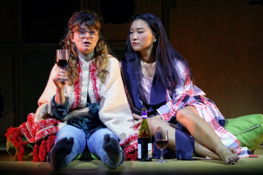 On stage, Amber McMahon and Michelle Lim Davidson sit close together drinking wine, McMahon looking shocked.