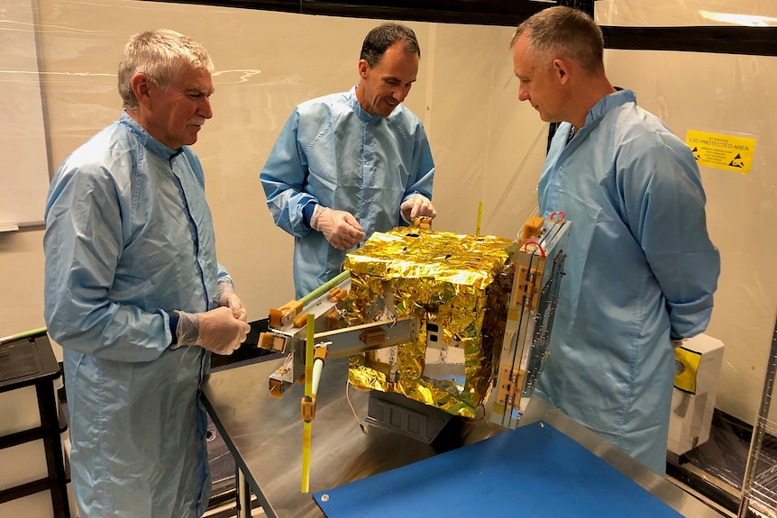 Three men look at a small satellite on a table.
