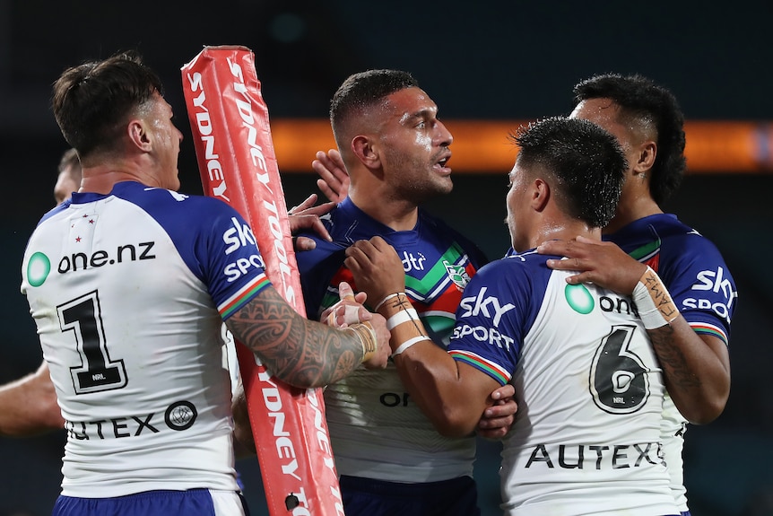 A group of NRL Warriors players gather around a teammate who has just scored near the corner flag.