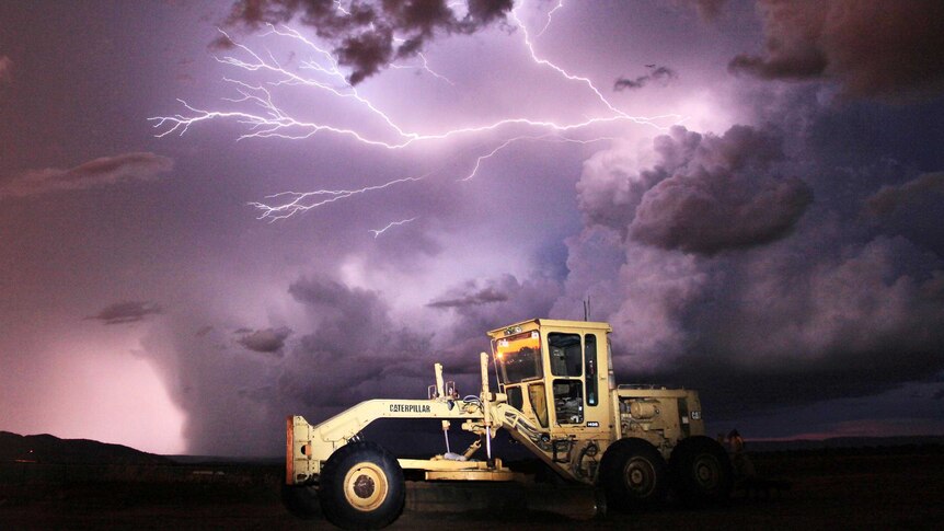 Lightning arcs across the sky over a grader sitting on a property during a night time storm.