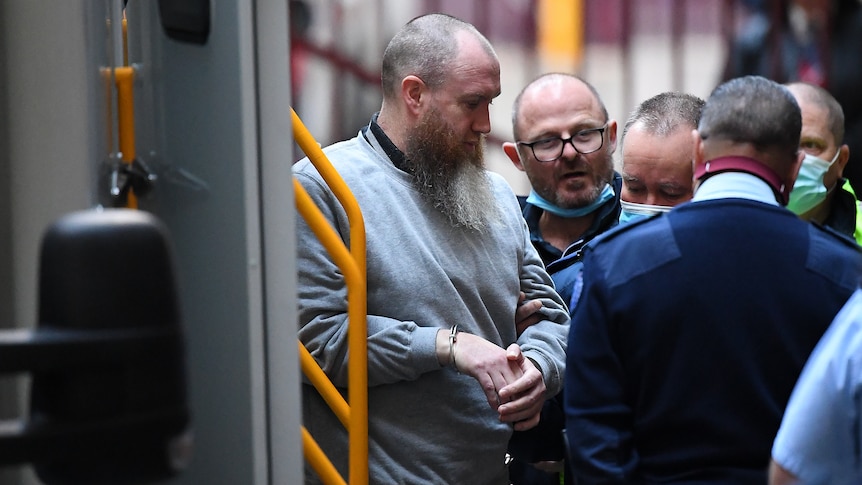 A handcuffed man with a long beard surrounded by guards as he enters court.