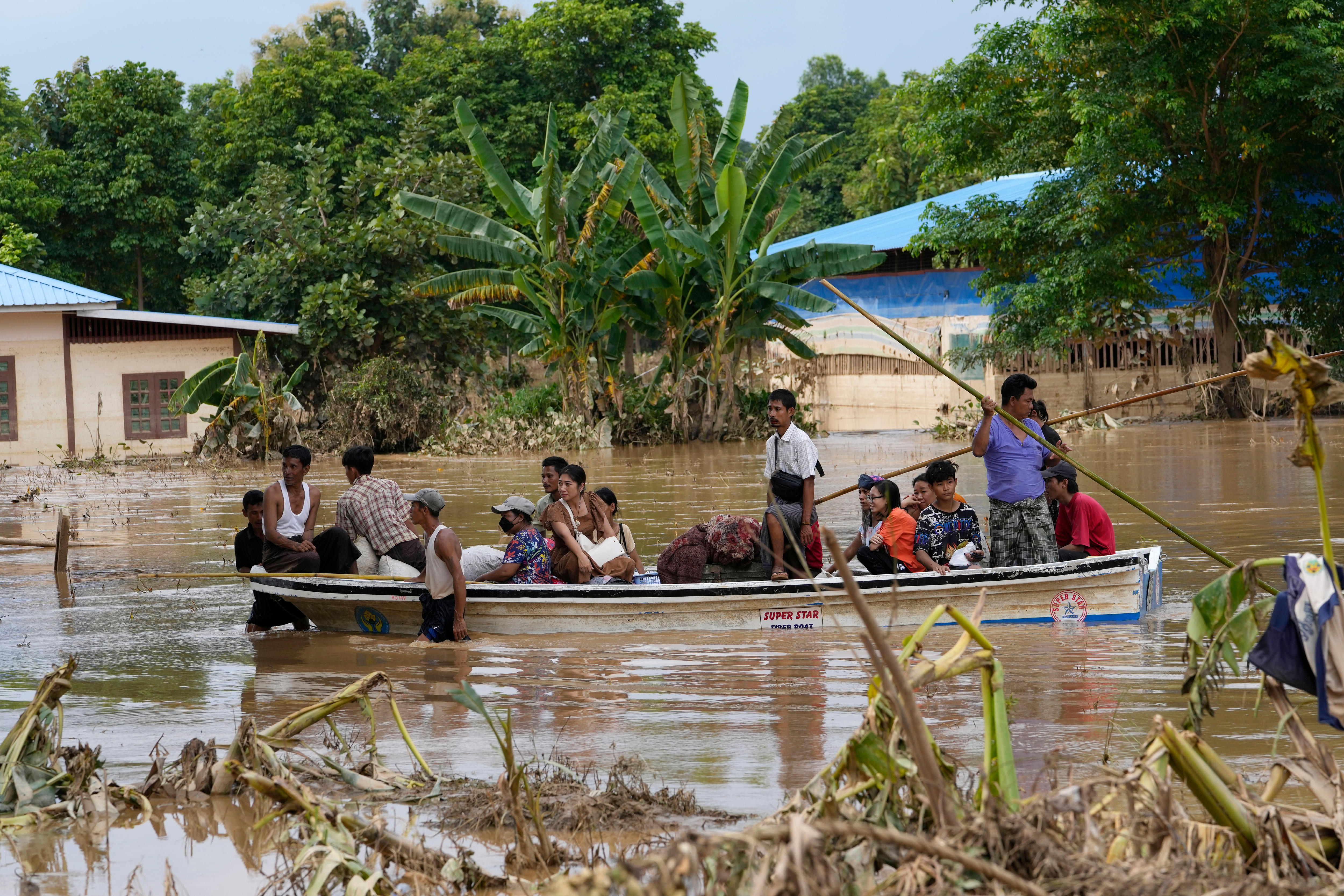 At least 16 people sit in a metal boat that is being pushed through floodwaters by two men using long wooden sticks