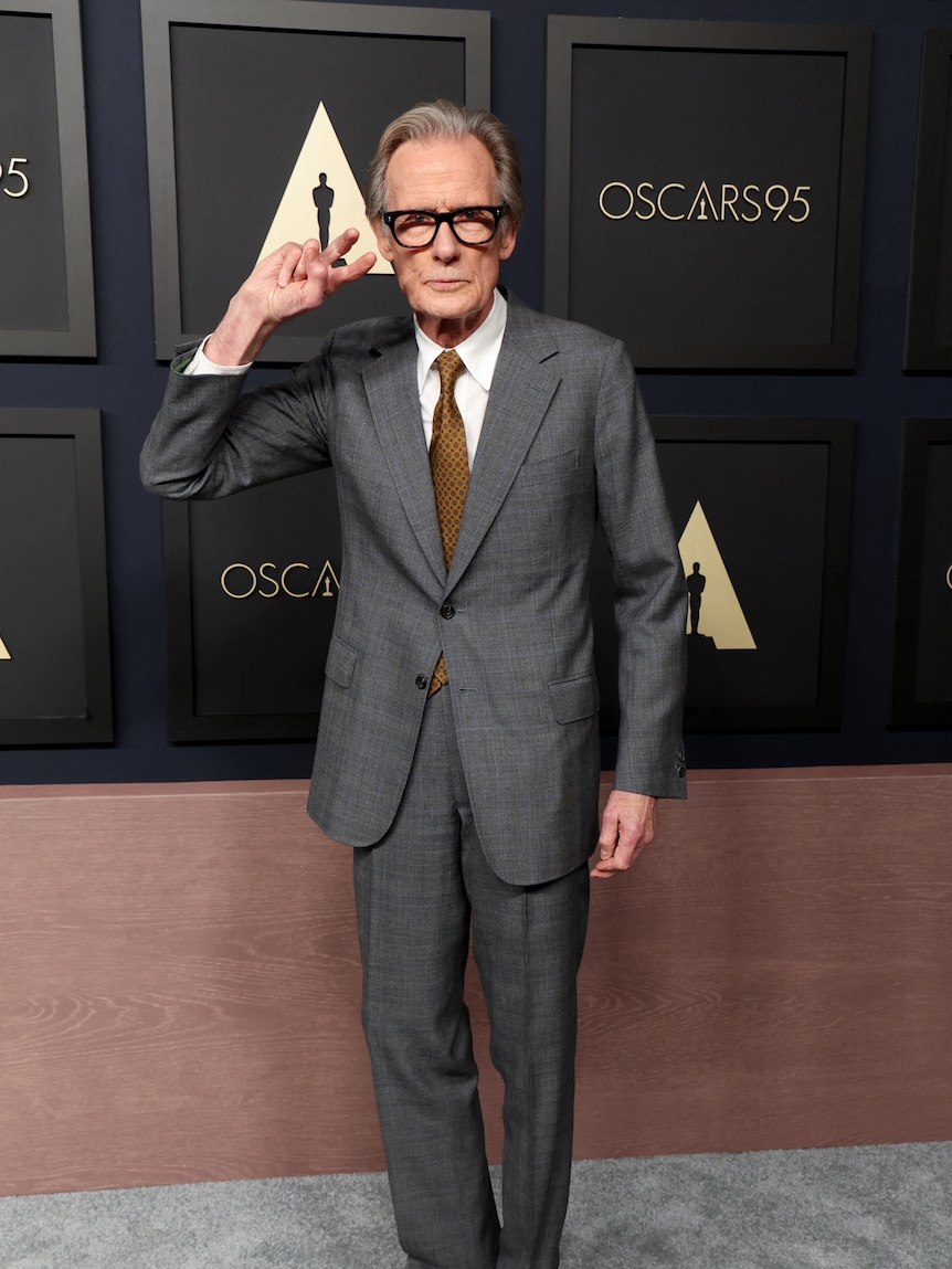 Bill Nighy holds a peace sign on the Oscars luncheon red carpet.