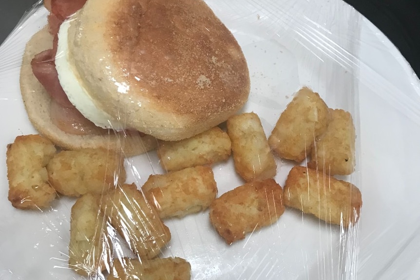  An egg and bacon roll and fried potatoes under cling wrap on a white plate