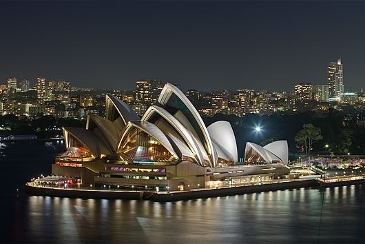 The iconic white sails of the Sydney Opera House, lit up at night with lights shining out over the harbour water.