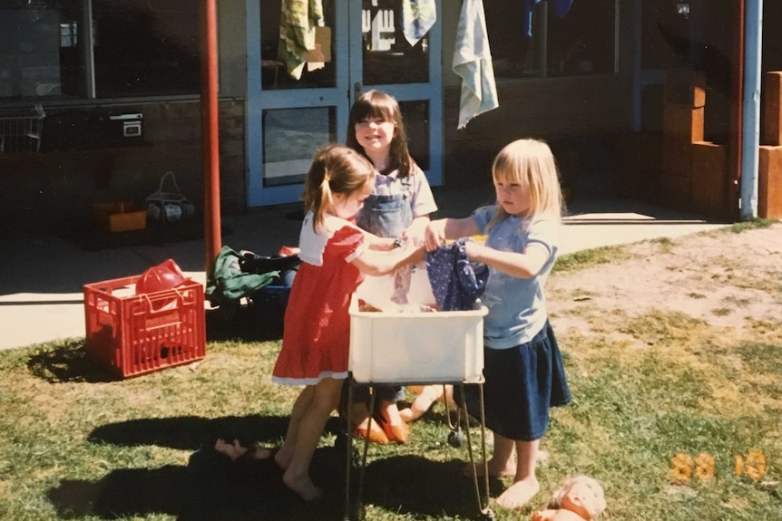 Three girls play with doll clothes around a make-shift wash tub.