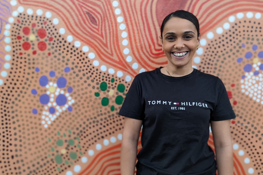 Rita smiles in a portrait in front of an Aboriginal dot painting mural