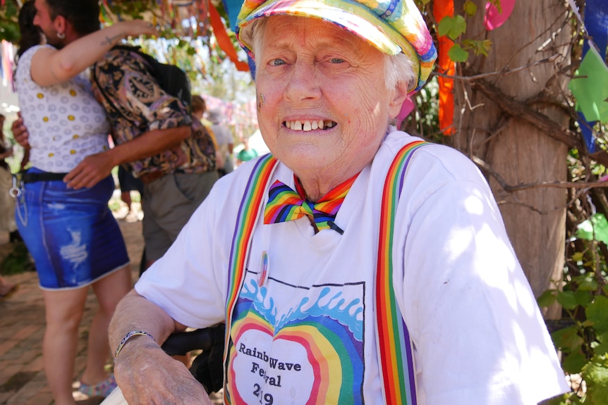 a older woman smiles at the camera wearing rainbow suspenders, a rainbow hat and white t-shirt with rainbow logo.