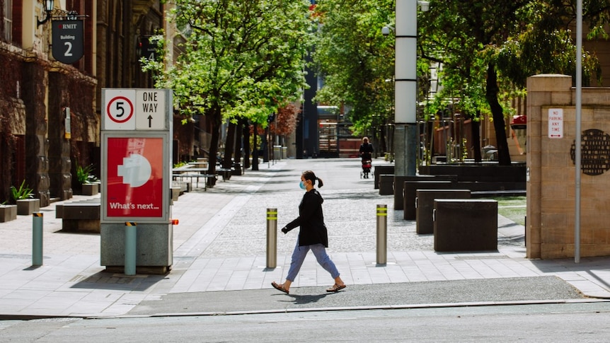 A woman wearing a blue surgical face mask is seen striding past an RMIT building on a sunny day in Melbourne.