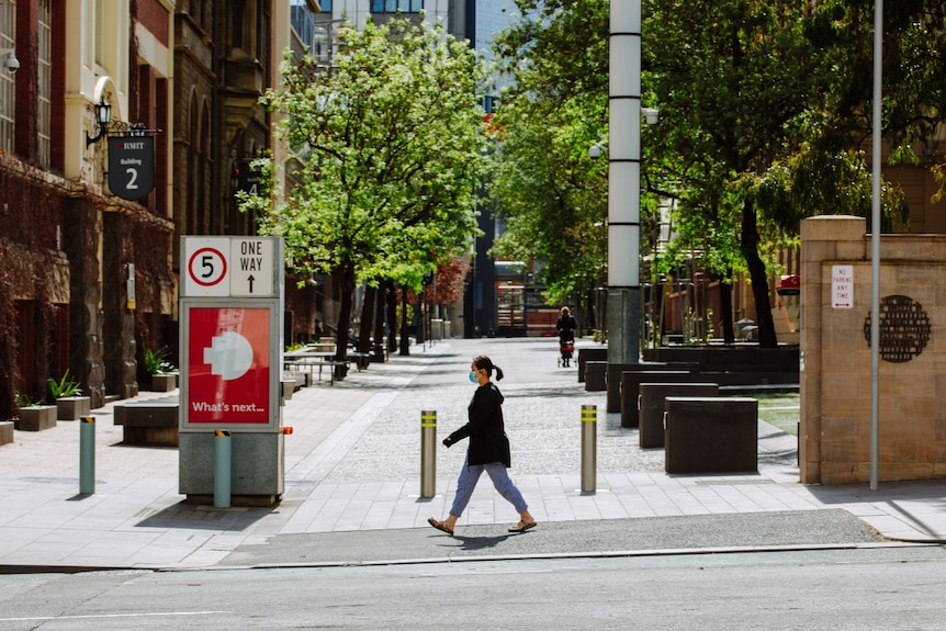 A woman wearing a blue surgical face mask is seen striding past an RMIT building on a sunny day in Melbourne.