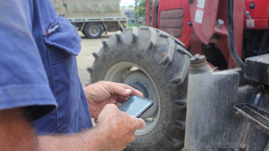 A man in a blue work shirt using a mobile phone, with a tractor in the background.