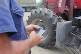 A man in a blue work shirt using a mobile phone, with a tractor in the background.