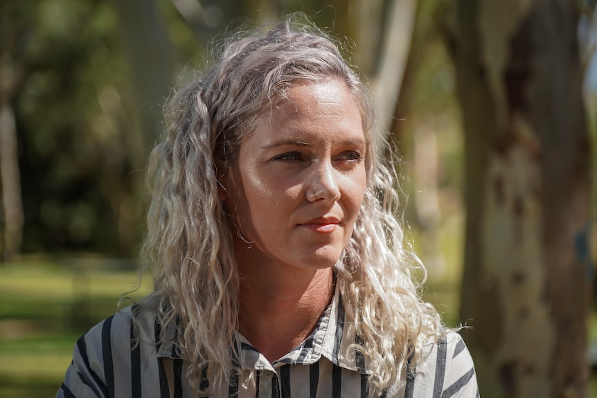 A woman with blond hair sits outside, gazing into the distance