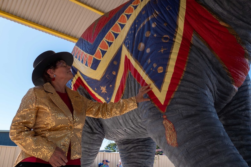 A woman in a gold sparkly jacket and a top hat touches and admires a colourful lifesize elephant statue.
