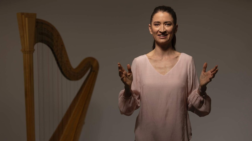 Harpist Genevieve Lang stands with her hands apart as if she's explaining something. A concert harp sits in the background.