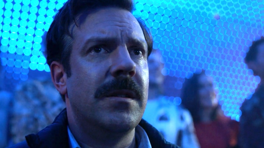 Man with intense, worried expression stands with people talking behind him in blue-lit room