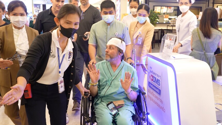 A man with his head bandaged and in a wheelchair being whisked away and surrounded by people