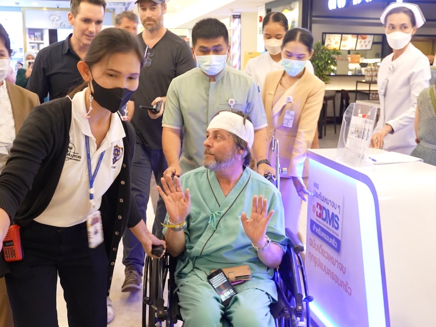 A man with his head bandaged and in a wheelchair being whisked away and surrounded by people
