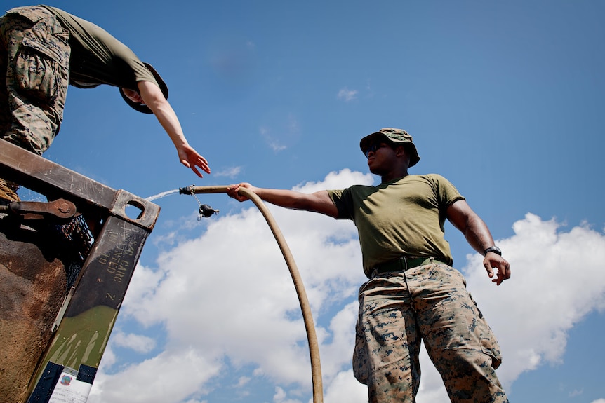 A US Marine in camo fatigues passing a hose up to a fellow marine standing on top of a platform.