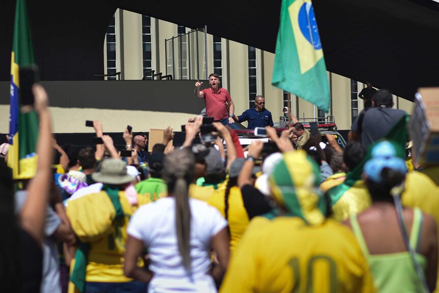A man angrily gestures from a raised platform as he addresses a crowd including people waving Brazilian flags