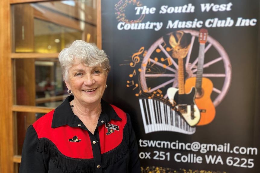 An older lady with grey hair stands in front of a sign with a guitar on it