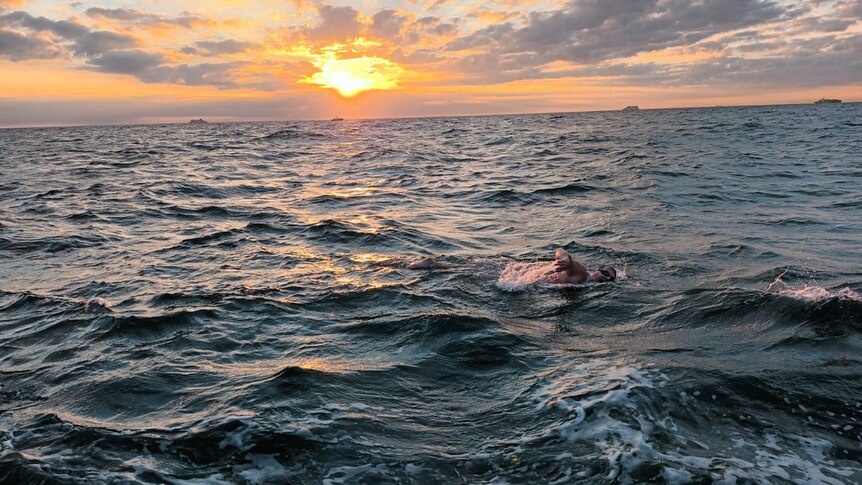 A man swims the English Channel. He is swimming free style his right arm out of the water. The sun is rising on the horizon. 