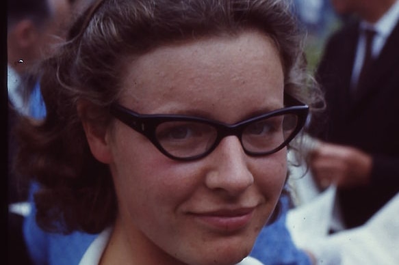 Photo of Jocelyn Bell (Burnell) in 1967. Young woman smiling at the camera with black framed glasses, white cardigan, blue top