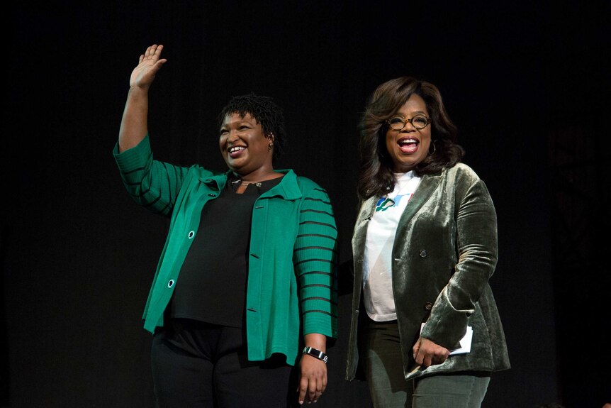Stacey Abrams smiling and waving next to Oprah Winfrey