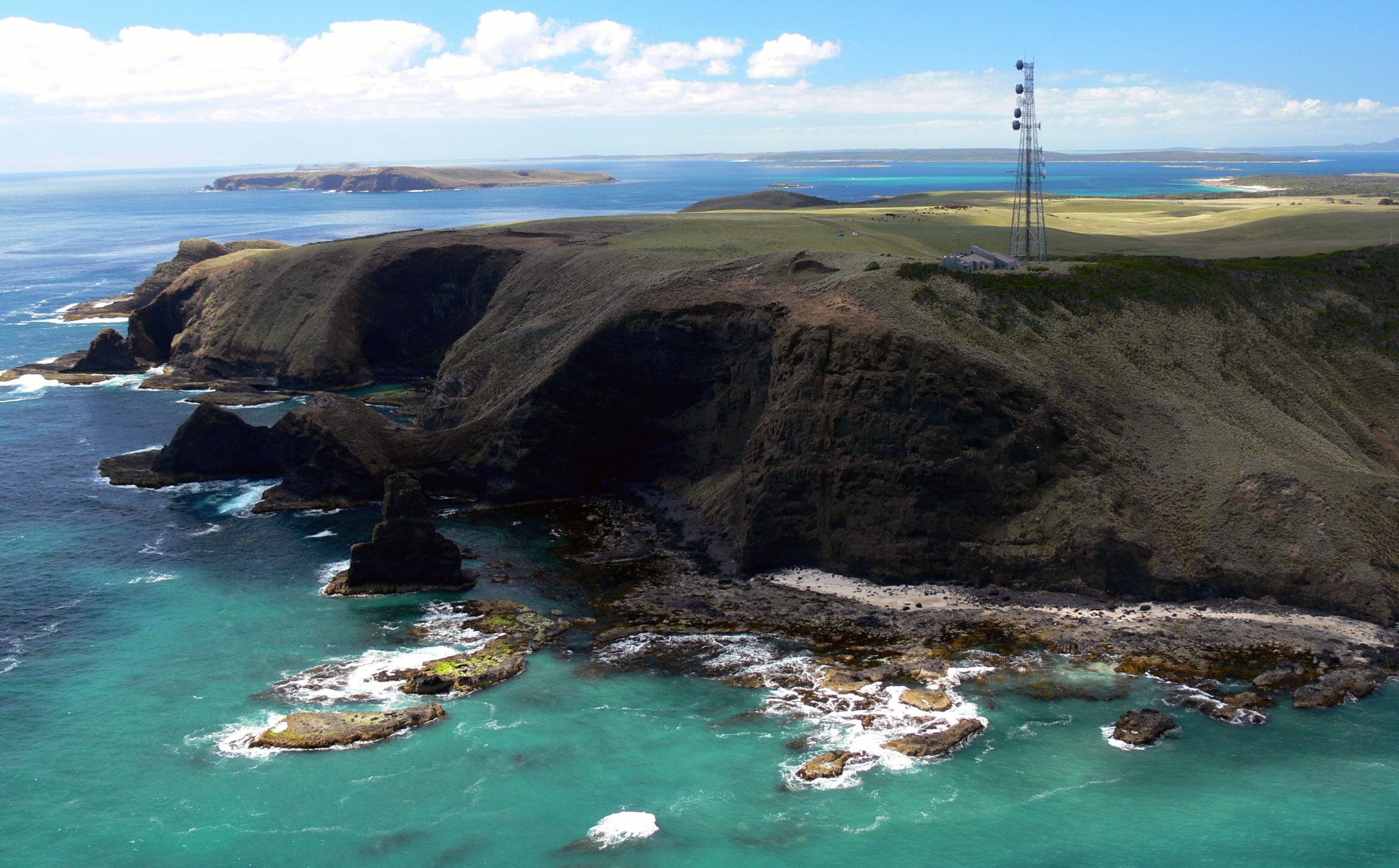 View of cliffs and green land mass with blue water in the foreground. There's an antenna on the edge of the land.