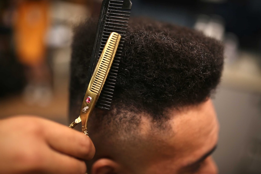A man with a hi-top fade hair style has his hair cut by a barber with gold coloured scissors.