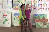 A dark-skinned lady with black hair stands next to a fair-skinned lady with brown hair in front of some posters of art.