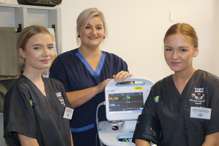 two students and one nurse standing next to a medical monitor