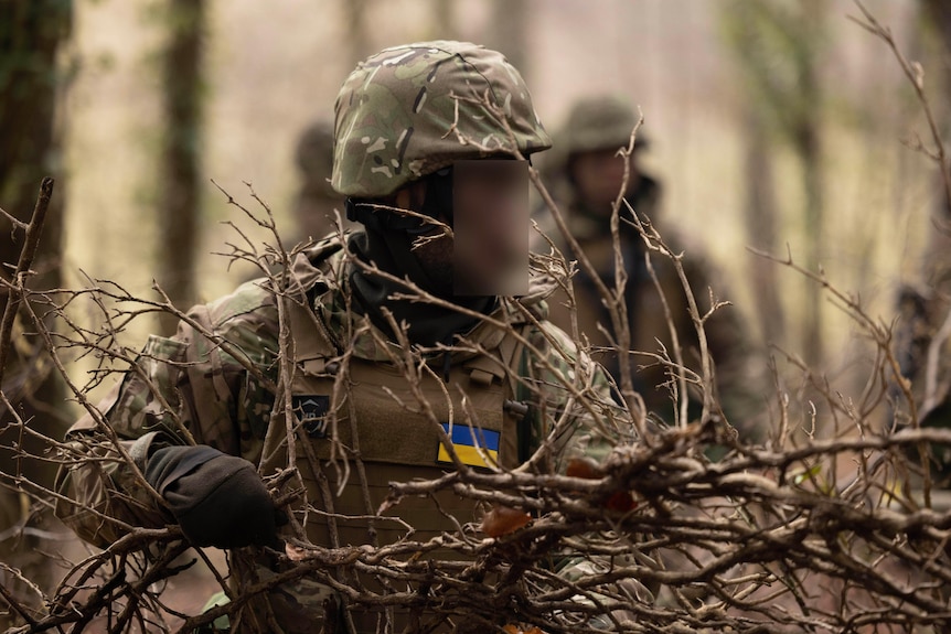 A soldier whose uniform is emblazoned with the Ukrainian flag carries a tree branch in a forest