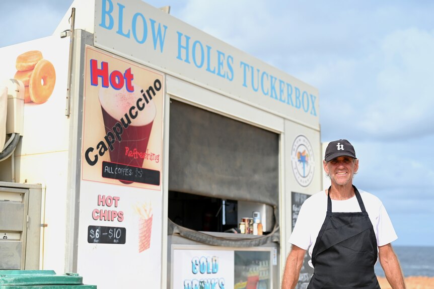 A man in an apron, white shirt and black hat standing in front of a food caravan at a sandy coastal area.
