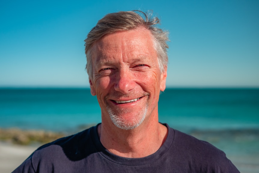 A man smiling to the camera with a beach faded in the background.