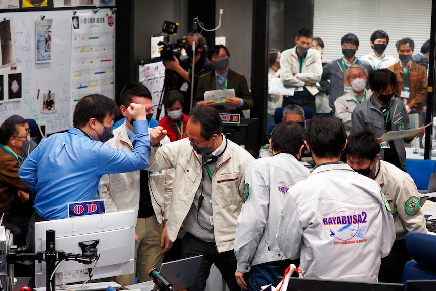 You view a crowd of Japanese men celebrating while wearing lab coats and face masks in an office.
