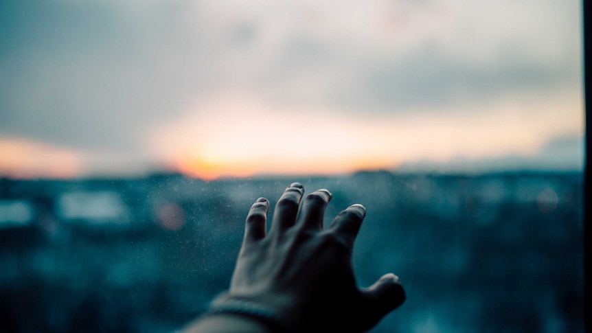A hand reaches towards a window showing cloudy skies.