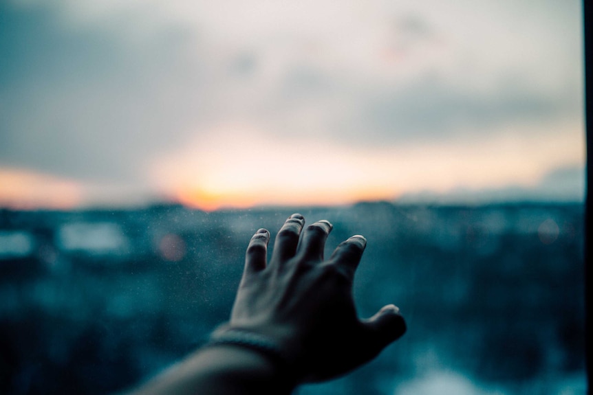 A hand reaches towards a window showing cloudy skies.