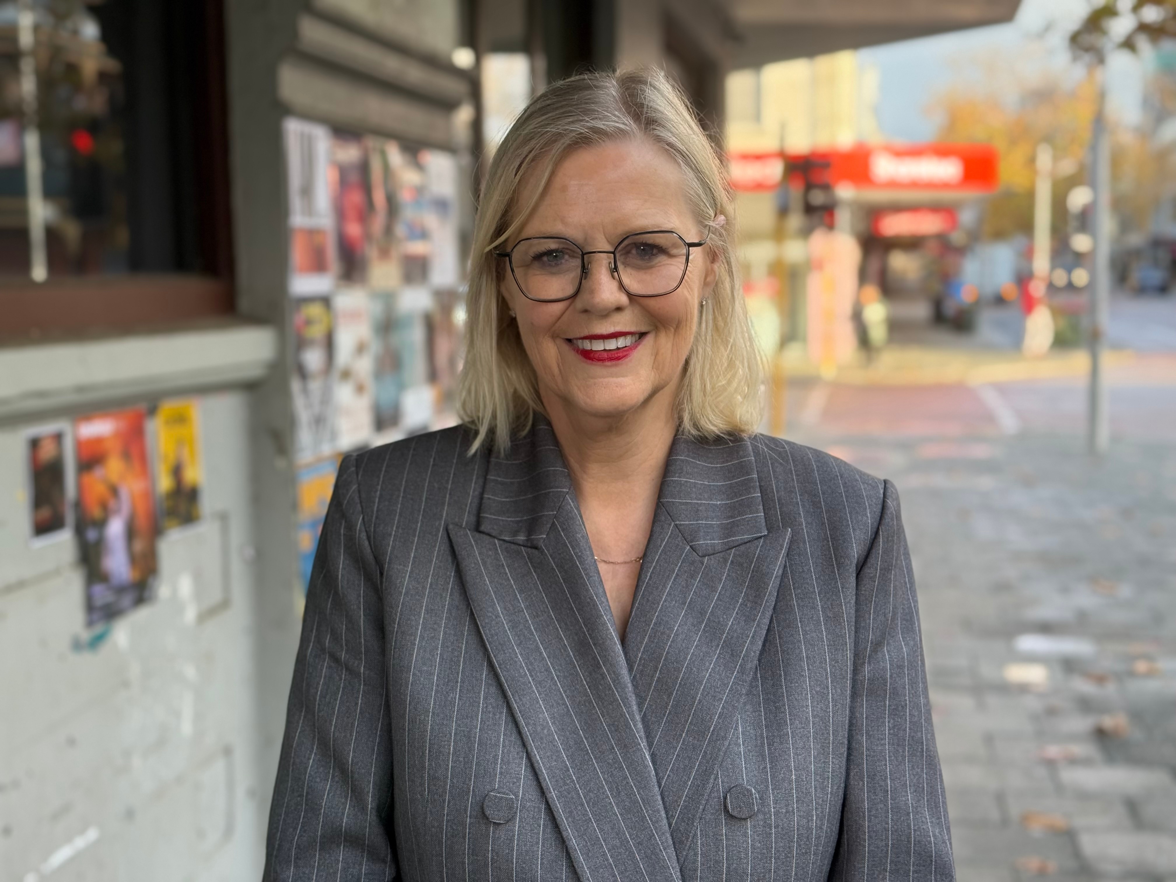 National Children's Commissioner Deb Tsorbaris smiles posing for a photo on a city footpath wearing a pinstriped grey jacket.