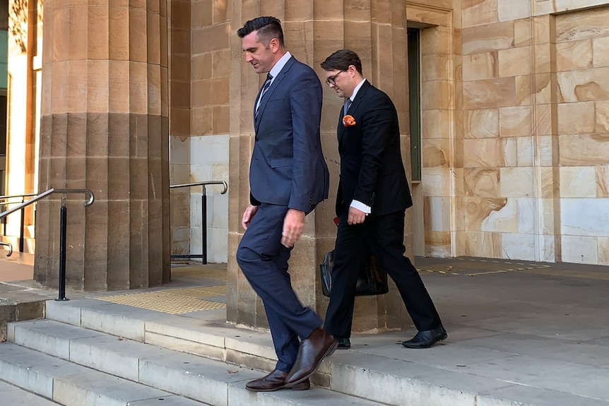 Two men in suits walk down stairs outside a sandstone building