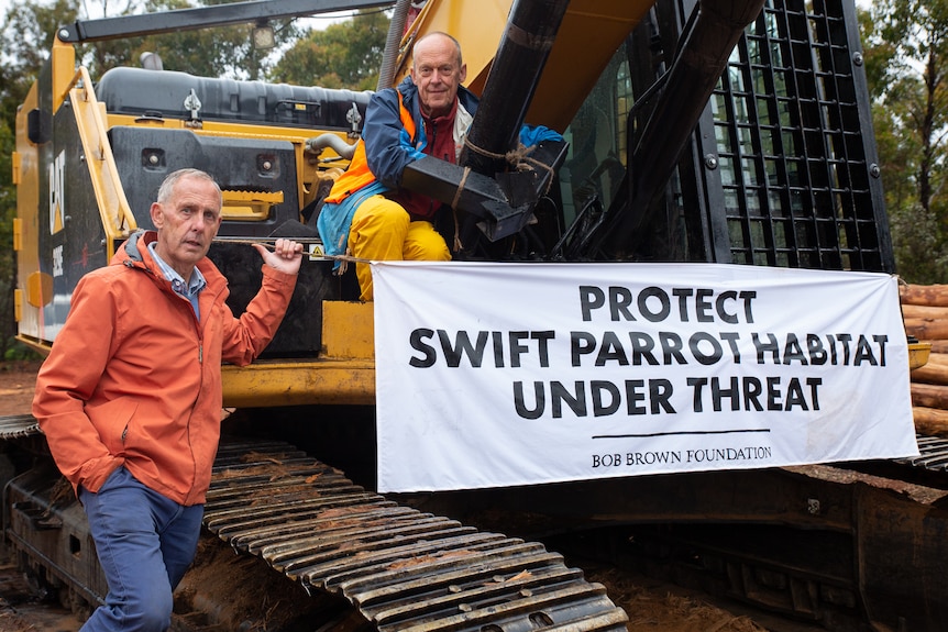 A man stands next to a large earth mover in the forest, as another man sits on top of it behind a protest sign