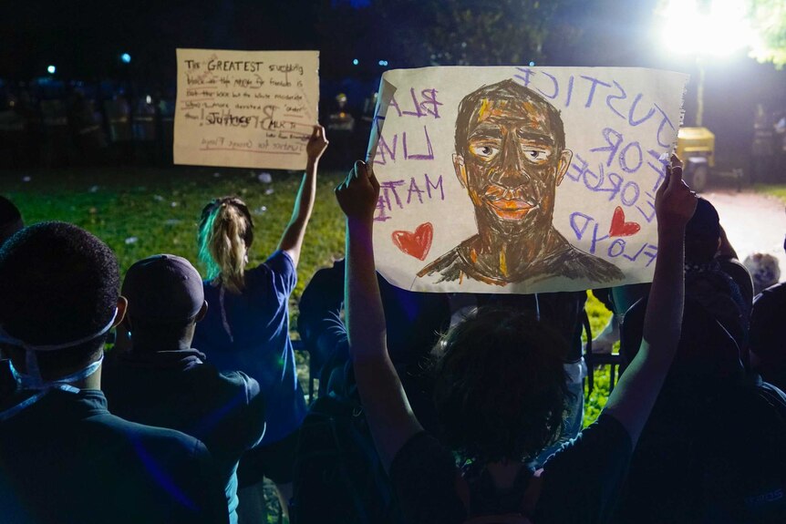 People standing in a park at night time hold up signs that say "Justice for George Floyd" and "Black Lives Matter"