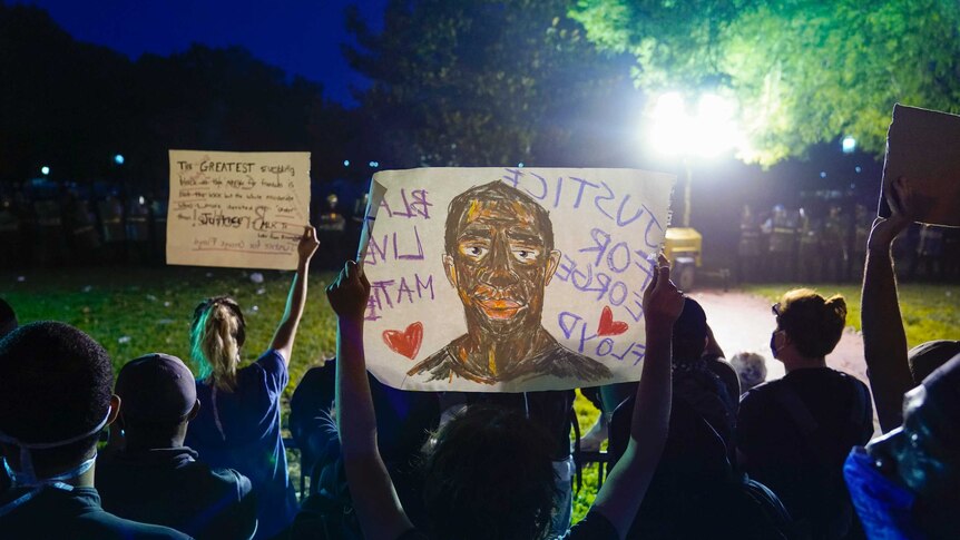 People standing in a park at night time hold up signs that say "Justice for George Floyd" and "Black Lives Matter"