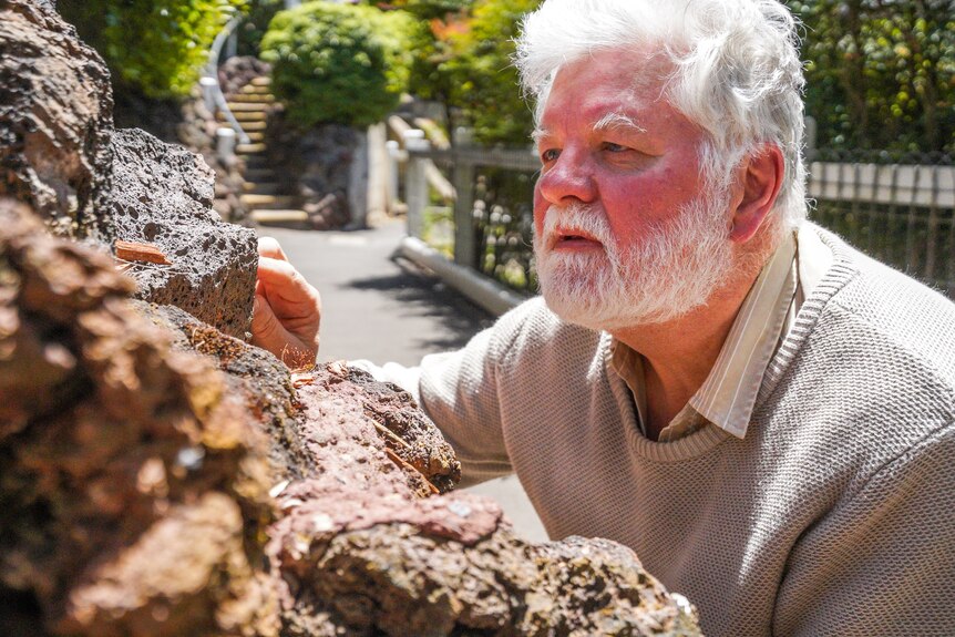 A man with white hair and a beard closely examines a rock wall.