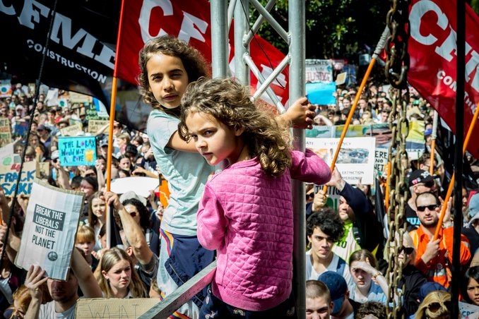 Two young girls climb an oil rig at a climate change protest. They are surrounded by protesters holding banners and signs.