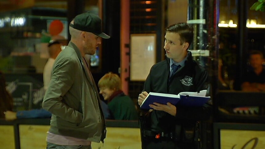A police officer takes notes while speaking to a man in a baseball cap.