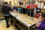 Pasta in containers on a commercial kitchen bench with people standing around holding lids.